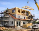 Construction d'une extension maison étage avec une grue qui installe une charpente en bois. Des ouvriers travaillent sur des échafaudages.