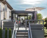 Terrasse avec mobilier de jardin et une véranda bois et alu. Parasol ouvert, escalier menant à la terrasse.