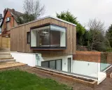 Une extension de maison moderne en bois avec une grande fenêtre en saillie. L'agrandissement maison s'intègre à un jardin en pente avec des murs de briques et des garde-corps en verre.
