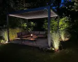 Une **pergola au milieu du jardin** illuminée la nuit, avec un salon de jardin confortable et des herbes hautes éclairées.