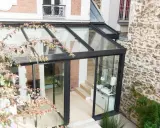 Une pergola transformée en véranda moderne, avec une structure en verre et métal, s'intégrant à une façade en pierre et brique.