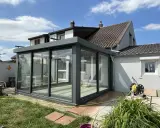 Une véranda moderne, conçue par un architecte, prolonge une maison. Elle est en verre et métal gris, avec un toit plat. Le jardin est verdoyant.