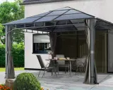 Une tonnelle def. avec table et chaises sur une terrasse pavée, devant une maison blanche et un jardin verdoyant.