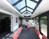Une extension veranda salle à manger moderne avec un toit vitré, une table noire et des chaises rouges, un canapé blanc et une vue sur le jardin.