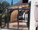 Une élégante veranda demi ronde en verre et métal, avec des stores rétractables, s'ouvre sur un jardin fleuri.