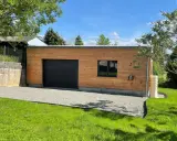 Extension garage bois accolé maison, avec porte noire, fenêtre et jardin verdoyant.