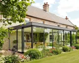 Une élégante veranda en verre s'adosse à une maison en pierre, offrant une vue sur une cuisine et une salle à manger. Le jardin fleuri entoure cette extension moderne.