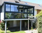 Moderne veranda adossée au mur, avec une chaise verte visible à travers les grandes baies vitrées. Le soleil brille sur le jardin verdoyant.