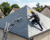 Un couvreur travaille sur un toit 4 pans, posant des ardoises. Une échelle et une scie circulaire sont visibles.