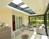 Une **veranda qui s'ouvre entièrement** sur un jardin verdoyant. Mobilier confortable, table et chaises pour profiter du soleil.