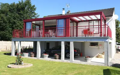 Une maison avec une veranda terrasse rouge vif, offrant une vue sur un jardin verdoyant et une voiture garée sous le balcon.