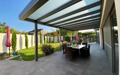 Une belle pergola moderne avec un toit vitré pour couvrir l'espace repas extérieur. Le jardin est bien entretenu, avec une pelouse verte et des cyprès élancés.