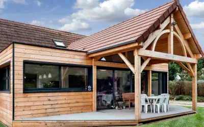 Extension de maison en bois avec terrasse sur dalle béton. Une table et des chaises sont installées sous un auvent.