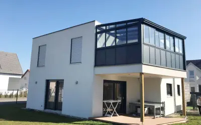 Maison moderne avec une veranda 2 niveaux vitrée, offrant une extension lumineuse. Terrasse en bois en contrebas.