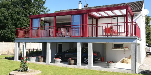 Une maison avec une veranda terrasse rouge vif, offrant une vue sur un jardin verdoyant et une voiture garée sous le balcon.