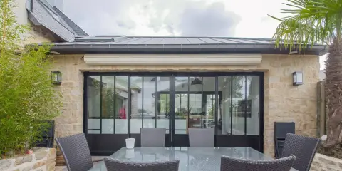 Terrasse avec mobilier autour d'une extension maison style atelier aux grandes verrières noires. Bambou et palmier apportent une touche végétale.