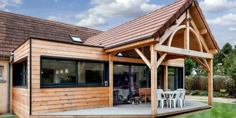Extension de maison en bois avec terrasse sur dalle béton. Une table et des chaises sont installées sous un auvent.