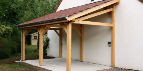 Un abri de voiture en bois avec une pente légère, adossé à une maison blanche.