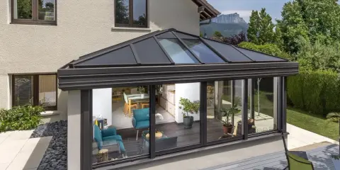 Une **pergola bioclimatique** moderne avec toit vitré, prolongeant une maison vers un jardin verdoyant. L'intérieur lumineux révèle un salon cosy et une salle à manger.