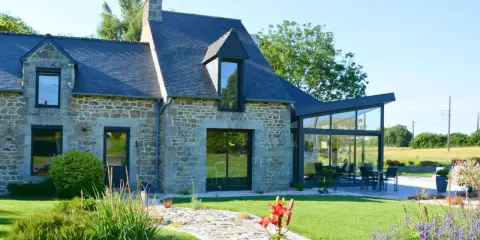 Maison en pierre avec une extension longere vitrée moderne. Un chemin pavé mène à la terrasse où une table est dressée.