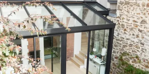 Une pergola transformée en véranda moderne, avec une structure en verre et métal, s'intégrant à une façade en pierre et brique.