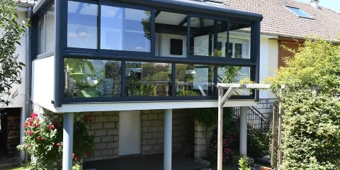 Moderne veranda adossée au mur, avec une chaise verte visible à travers les grandes baies vitrées. Le soleil brille sur le jardin verdoyant.