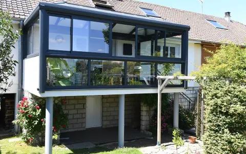 Une extension véranda toit plat moderne, avec de larges baies vitrées, s'intègre parfaitement à la maison. Un fauteuil vert est visible à l'intérieur.