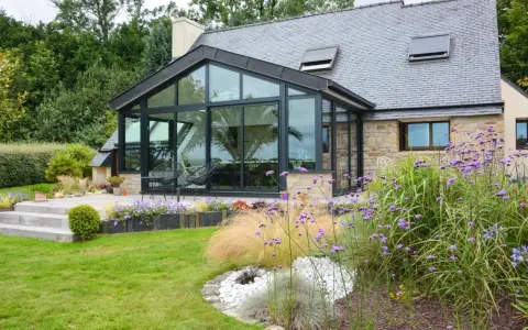 Maison avec extension toit vitrée, terrasse en bois et jardin fleuri.