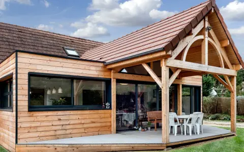 Extension de maison en bois avec terrasse sur dalle béton. Une table et des chaises sont installées sous un auvent.