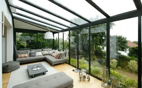 Salon lumineux avec canapé d'angle gris, table basse et vue sur jardin luxuriant. Idéal pour agrandir veranda et profiter de la nature.