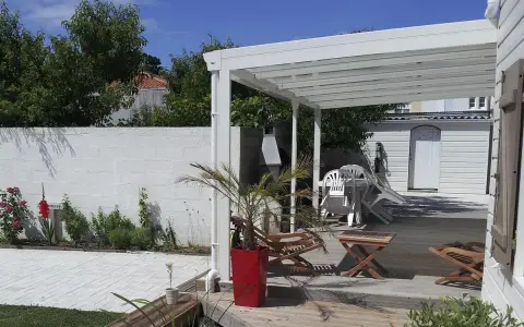 Terrasse en bois sous un préau semi-fermé, avec chaises empilées, une table, un palmier en pot et un barbecue.