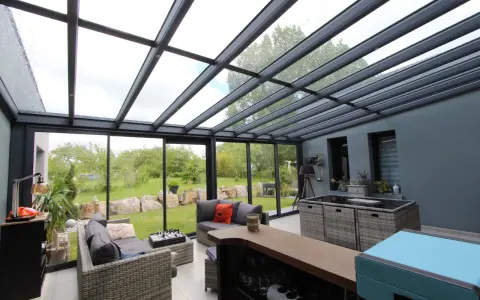Salon moderne avec un toit en verre laissant entrer la lumière. Des canapés confortables invitent à la détente, tandis que le jardin verdoyant offre une vue paisible.