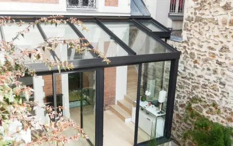 Une véranda bioclimatique moderne, avec une structure en verre et métal noir, s'intègre à une maison en briques et pierre. Avis positif sur son esthétique.