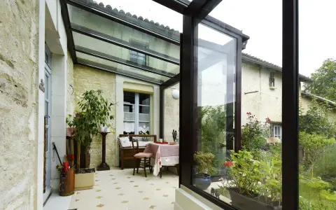 Une veranda avec toiture tuile, aménagée avec une table et des plantes, s'ouvre sur un jardin verdoyant.