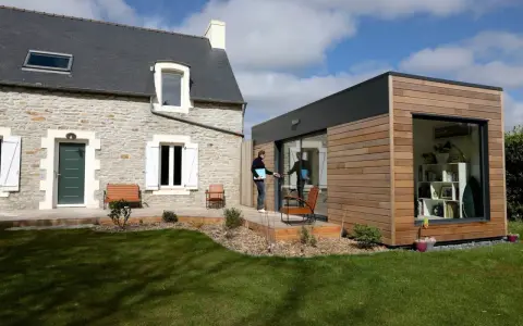 Une maison en pierre avec une extension moderne en bois. Un homme sort de l'extension, prêt à profiter du jardin.