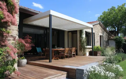 Construction d'une pergola moderne sur une terrasse en bois, avec une table et des chaises, devant une maison en pierre.