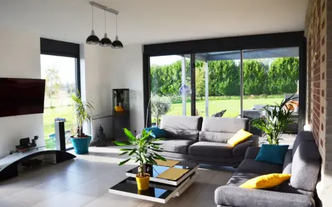 Salon lumineux avec canapés gris, table basse moderne et grandes baies vitrées donnant sur un jardin verdoyant. Une annexe maison parfaite pour se détendre.