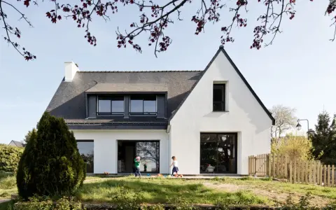 Une extension maison néo-bretonne moderne avec de grandes baies vitrées. Deux enfants jouent dans le jardin verdoyant devant la maison.