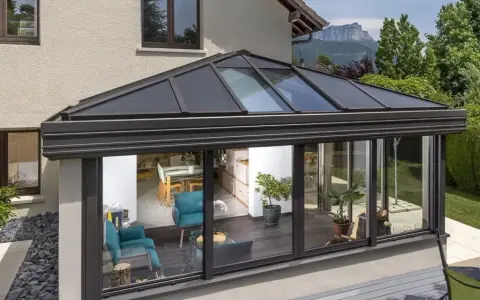 Une **pergola bioclimatique** moderne avec toit vitré, prolongeant une maison vers un jardin verdoyant. L'intérieur lumineux révèle un salon cosy et une salle à manger.