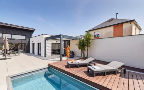 Une femme en robe colorée se tient près d'une pergola moderne, profitant de l'étanchéité pergola mur. Piscine et chaises longues complètent ce paysage de détente.