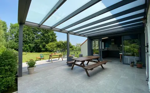 Terrasse avec table en bois sous une pergola bioclimatique. La hauteur de la structure permet de profiter du jardin verdoyant.