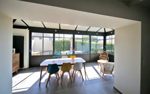 Salle à manger lumineuse dans une véranda chauffée, avec une table en bois et des chaises colorées. Un espace de vie agréable.
