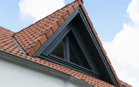 Un puit de lumière triangulaire s'intègre dans un toit en tuiles rouges, sous un ciel bleu et nuageux.