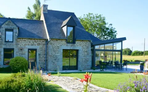 Maison en pierre avec une extension longere vitrée moderne. Un chemin pavé mène à la terrasse où une table est dressée.