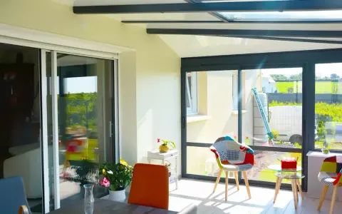 Intérieur lumineux d'une véranda avec un grand vitrage offrant une vue sur un jardin verdoyant. Des chaises colorées invitent à la détente.