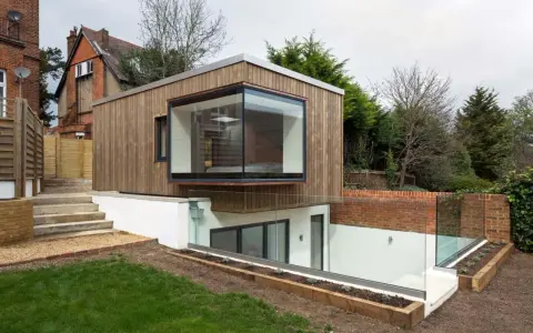 Une extension de maison moderne en bois avec une grande fenêtre en saillie. L'agrandissement maison s'intègre à un jardin en pente avec des murs de briques et des garde-corps en verre.