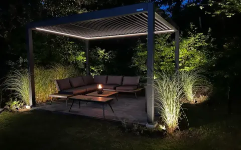 Une **pergola au milieu du jardin** illuminée la nuit, avec un salon de jardin confortable et des herbes hautes éclairées.
