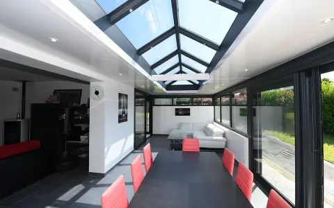 Une extension veranda salle à manger moderne avec un toit vitré, une table noire et des chaises rouges, un canapé blanc et une vue sur le jardin.