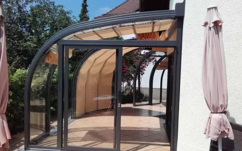 Une élégante veranda demi ronde en verre et métal, avec des stores rétractables, s'ouvre sur un jardin fleuri.