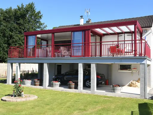 Une maison avec une veranda terrasse rouge vif, offrant une vue sur un jardin verdoyant et une voiture garée sous le balcon.